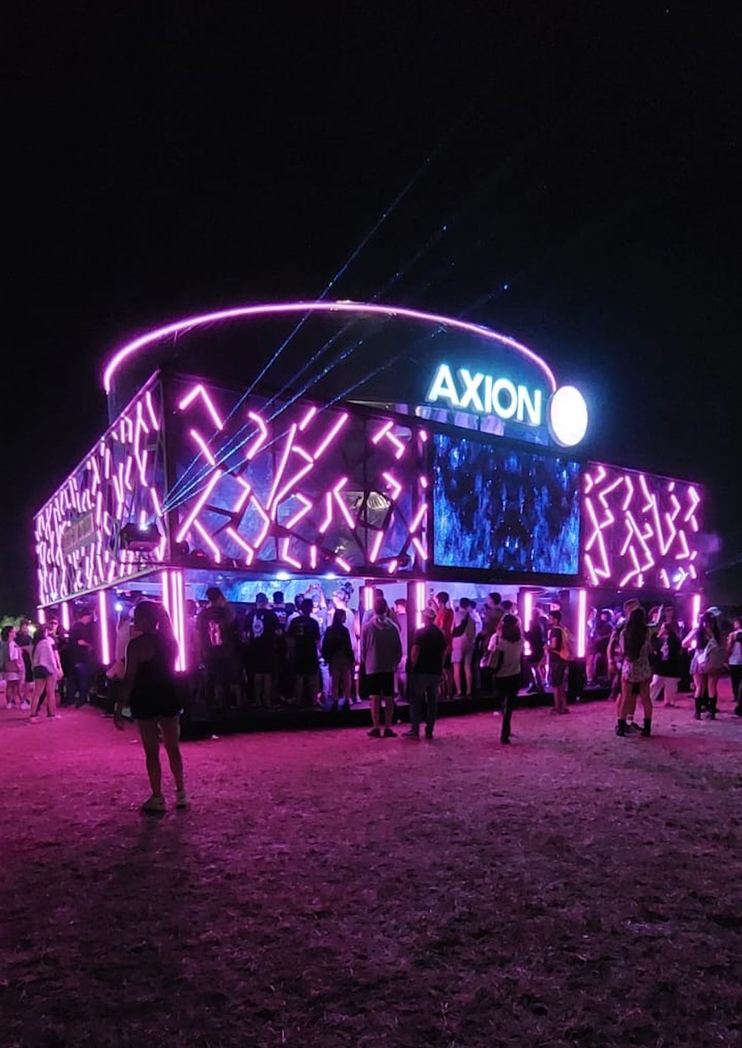 Stand de Axion de noche, con la estructura calada en luz rosa y el público adentro