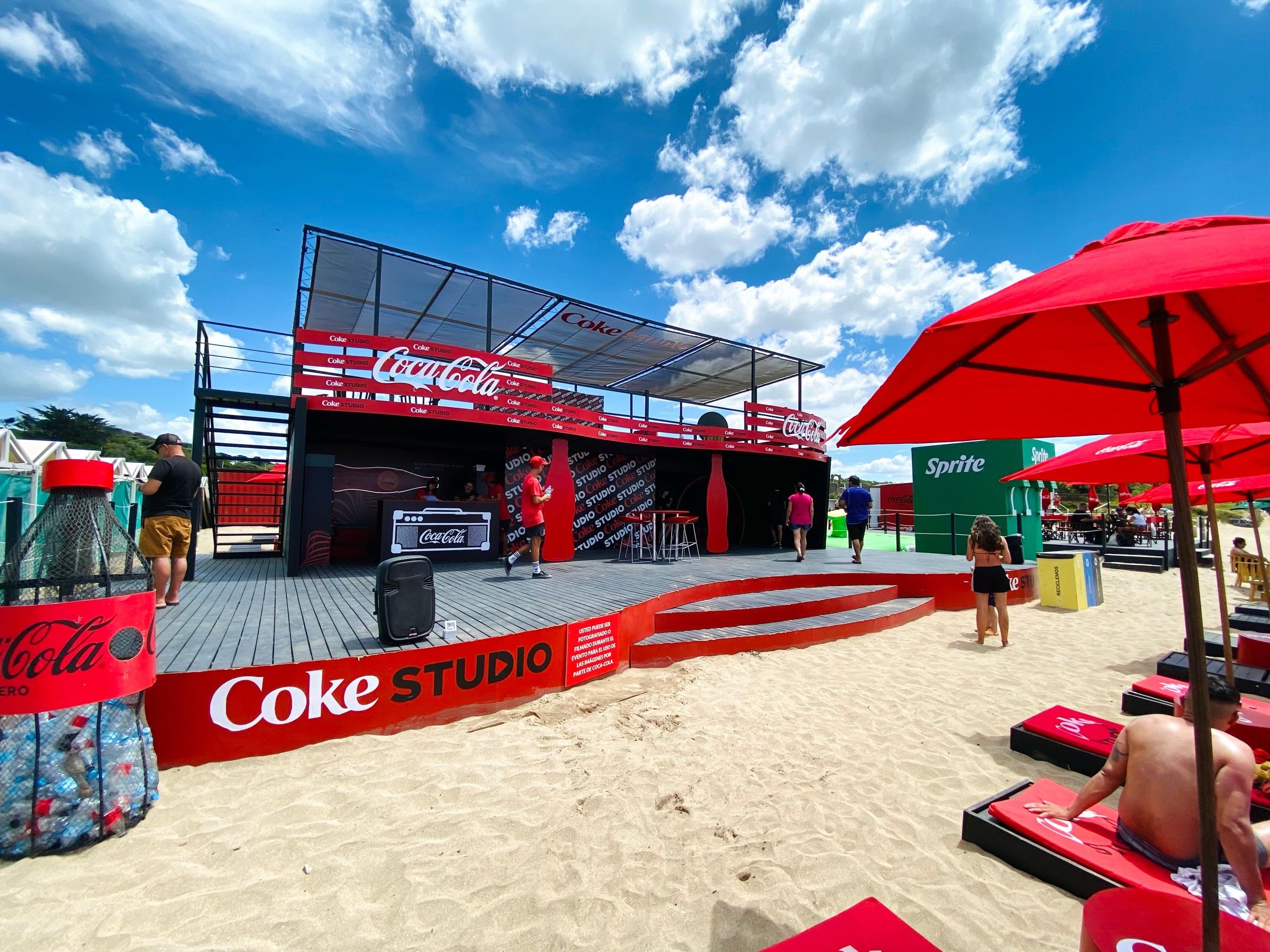Parador de Coca-Cola de dos plantas sobre la arena, con deck rojo y promotoras